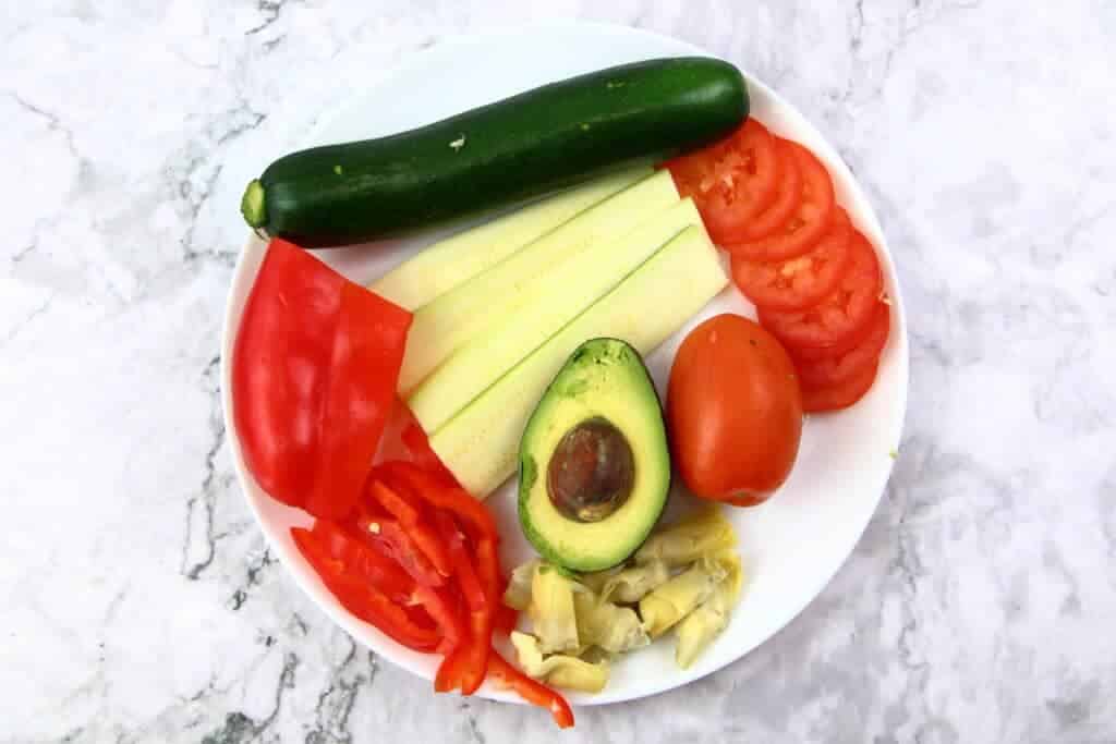 raw vegetables on a plate. Zucchini, avocado, tomatoes, red belle pepper, and artichoke.