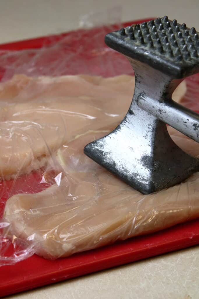 Chicken being hammered with a meat tenderizer between two pieces of plastic wrap