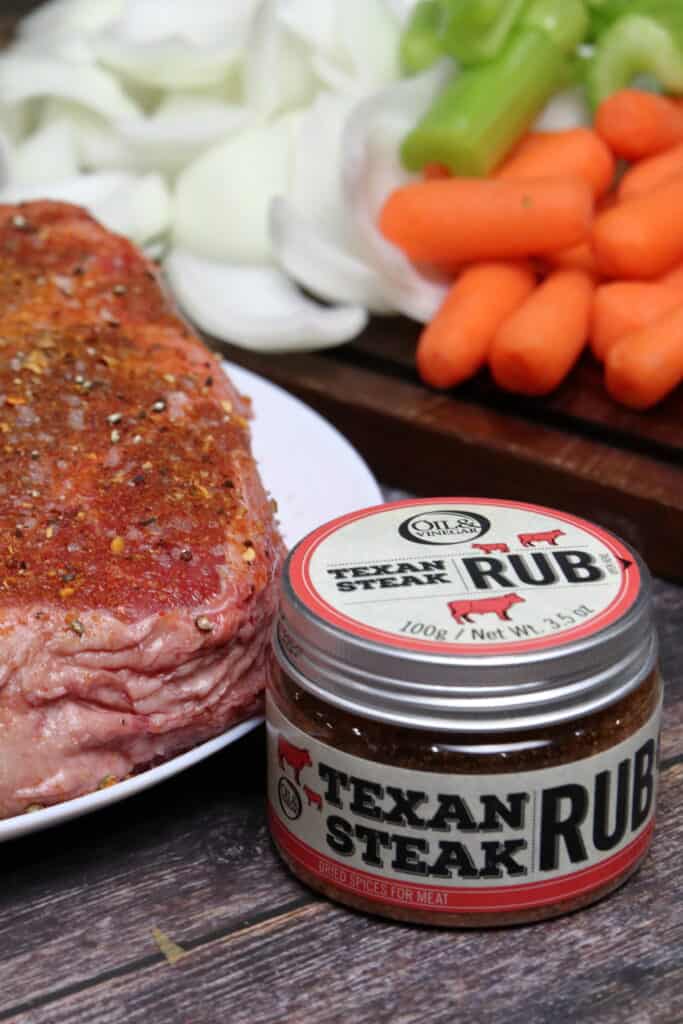 Seasoned chuck roast with veggie in the background next to a container of texan steak rub.