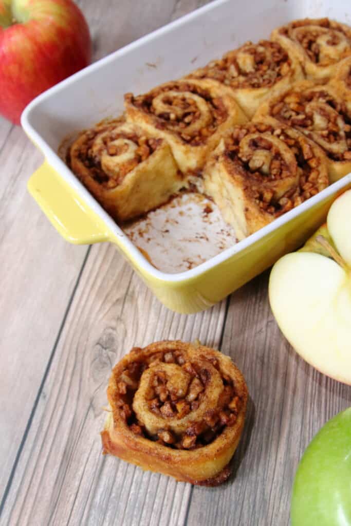 Pan of apple pie cinnamon rolls with one removed and placed on a table.